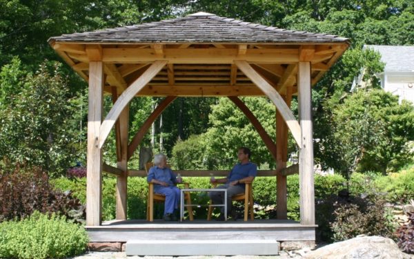 Two people sit under a timber frame gazebo in a garden in Massachusetts