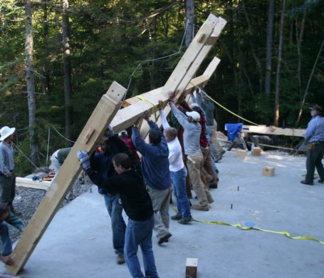 A group of people raise a section of a timber frame building into the air