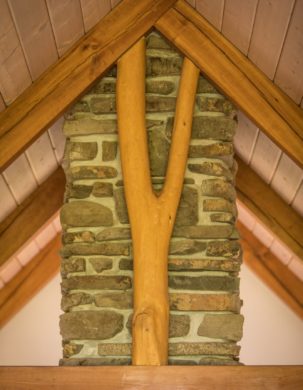 A forked timber scribed to rafters in front of a stone chimney in a timber frame with straw clay wall