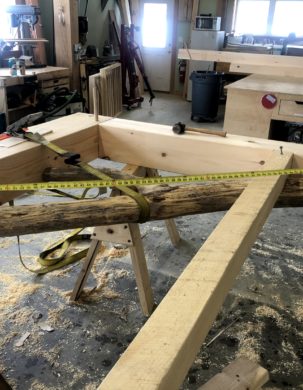 Inside a timber frame shop, a post, beam and forked brace are test fit and pegged together.