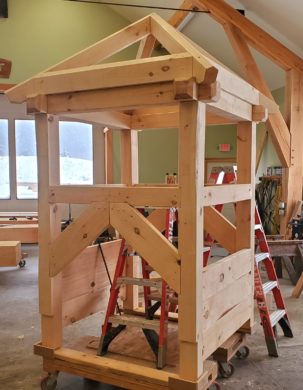 An image showing the cupola timber frame, without it's sheathing, decking, or windows. It sits on a rolling cart in a spacious woodshop.