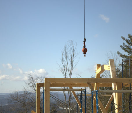 Two canted purlin posts are visible as the crane ball hangs just above the timber frame on a sunny Vermont morning