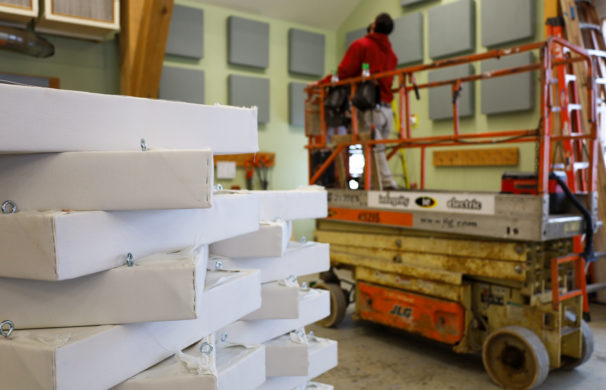 A craftsperson operates a scissor lift in the background, with a pile of sound dampening panels nearby